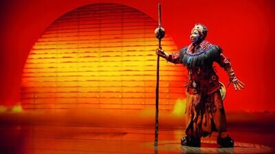 Der Musicaldarsteller von Rafiki aus Der König der Löwen steht auf der Bühne und hält einen Stab