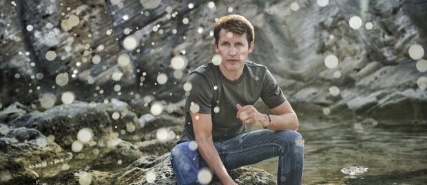 James Blunt sitzt auf einem Felsen am Wasser und schaut nach vorn