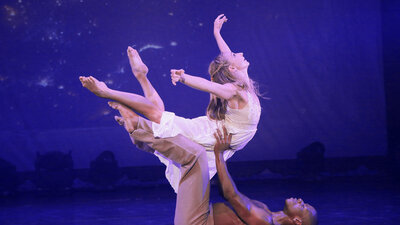 Ein Ballettänzer liegt auf dem Rücken auf einer Bühne und hebt eine Ballettänzerin hoch