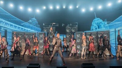 Die Darsteller des Musicals We Will Rock You stehen nebeneinander auf der Bühne und recken ihre rechten Arme nach oben