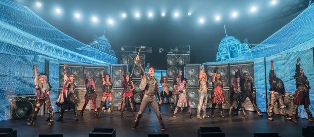 Die Darsteller des Musicals We Will Rock You stehen nebeneinander auf der Bühne und recken ihre rechten Arme nach oben