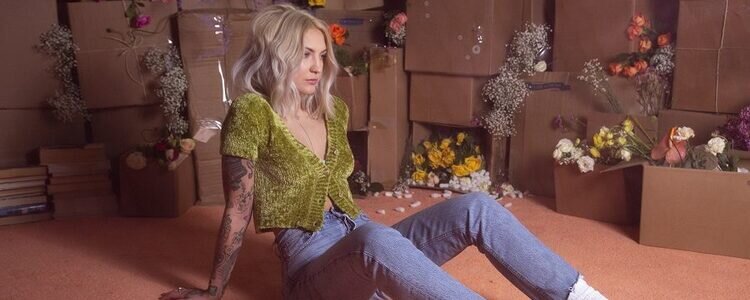 Julia Michaels sitzt auf dem Boden und schaut in die Ferne während im Hintergrund Kartons gefüllt mit Blumen stehen.  