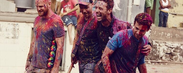 Die Mitglieder der Band Coldplay sind mit bunten Farben bekleckst und liegen sich lachend in den Armen.