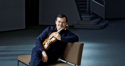 Frank Peter Zimmermann sitzt auf einer gepolsterten Bank mit einer Violine im Arm